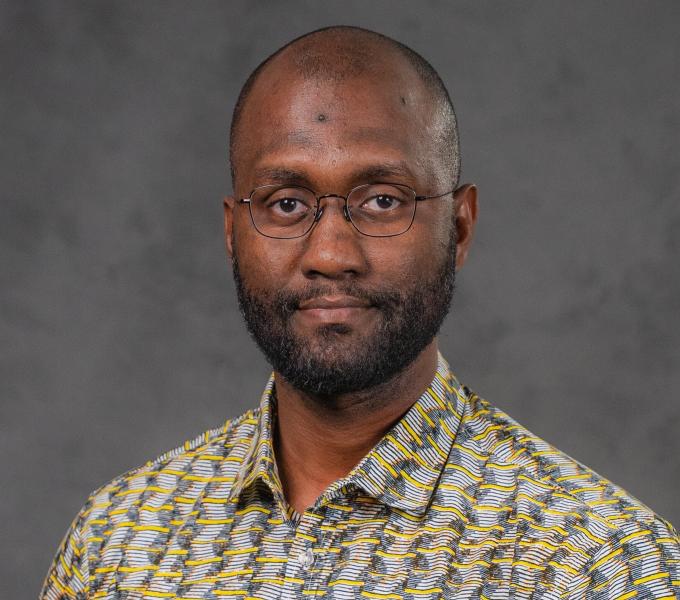 Headshot of a man in wire glasses and a bright yellow, gray, and white patterned shirt.