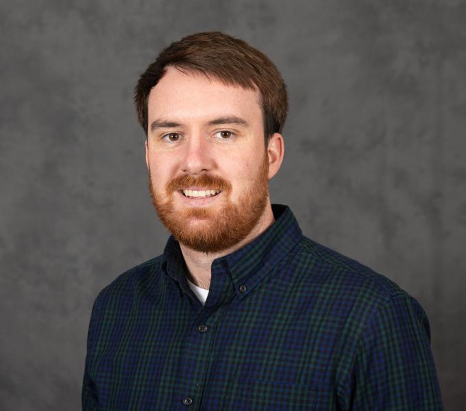 Headshot of a smiling bearded man in a dark navy and green plaid shirt.