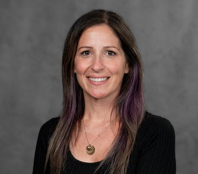 Headshot of a smiling woman with brown and purple hair in a black shirt and gold pendant.