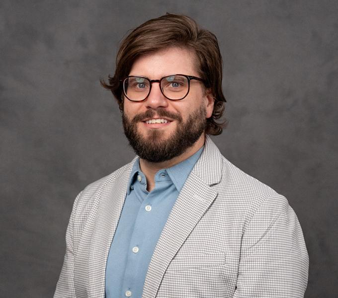 A man with long dark hair and a beard and glasses smiling in a blue shirt with a grey jacket.