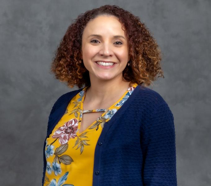 Headshot of smiling woman with honey-brown curly short hair in a yellow shirt and navy blue cardigan.