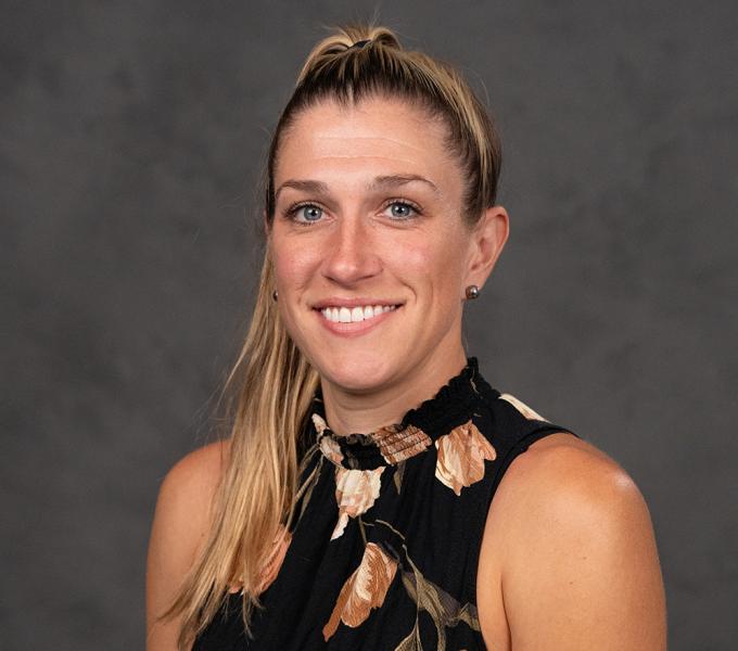 Blonde woman smiling in a floral sleeveless top in front of a grey background.