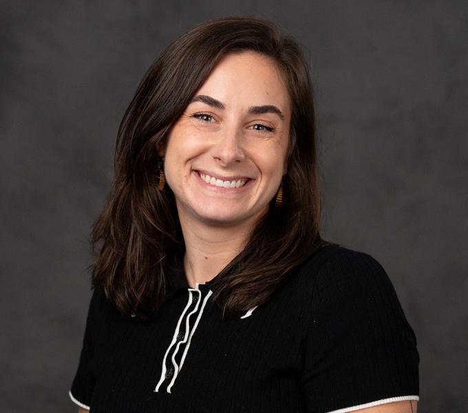 A woman with dark hair smiling in a black polo shirt in front of a grey background.