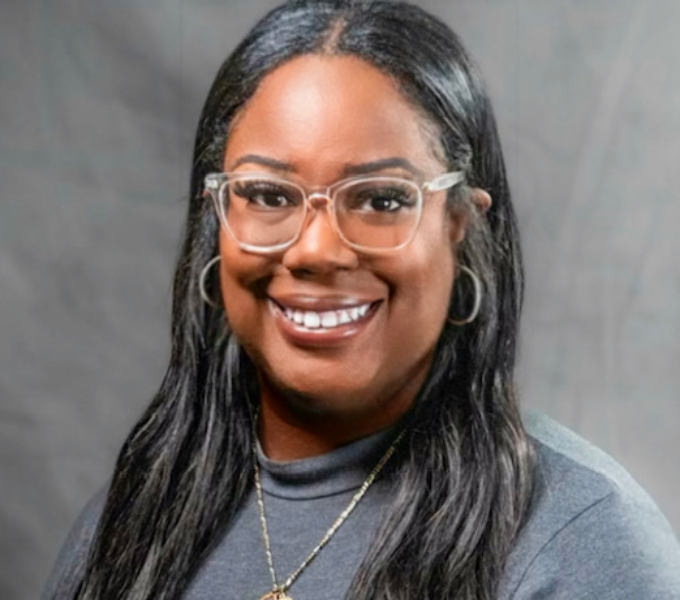 Headshot of a smiling woman in clear plastic glasses, a gold necklace, and a gray sweater.