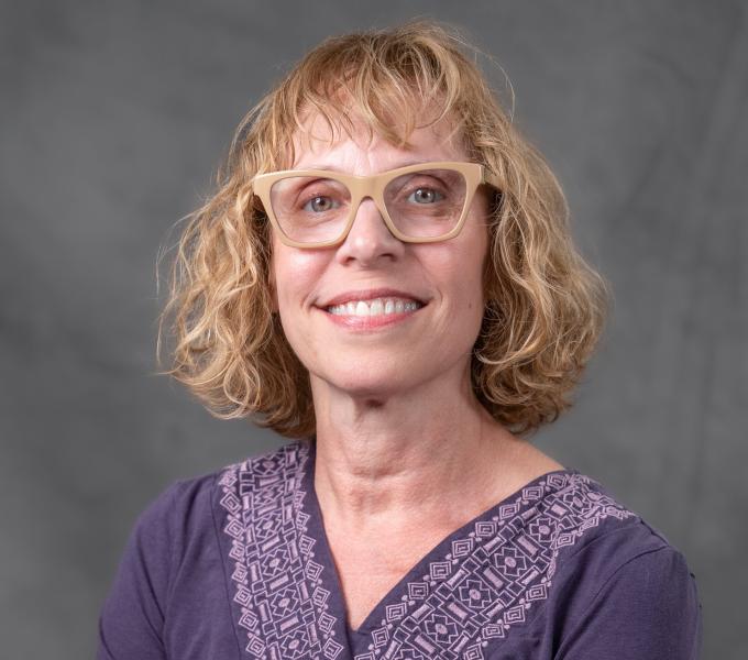 Headshot of a smiling woman in a purple shirt and tan glasses with short blonde hair.