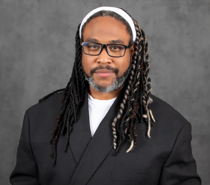 Headshot of a smiling man in a charcoal blazer, black glasses, and white headband.