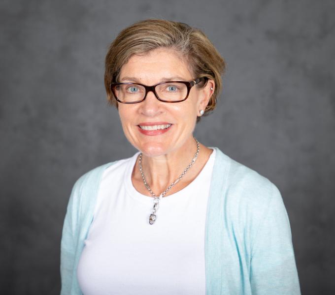 Headshot of smiling woman with short blonde hair, black plastic glasses, a white shirt and teal cardigan.