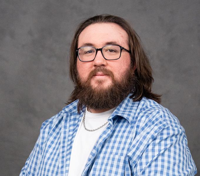 A man with long dark hair, a beard and glasses in a white shirt with a blue button down shirt.