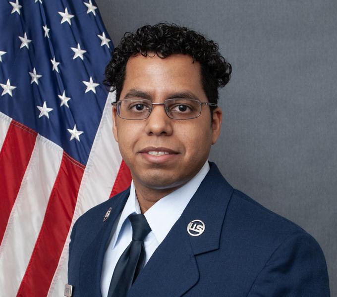 A man with short dark curly hair and glasses smiling in a navy suit.