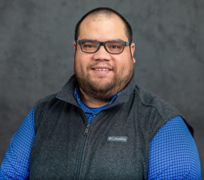 Headshot of smiling man in blue long sleeve shirt, gray vest, and black plastic glasses.