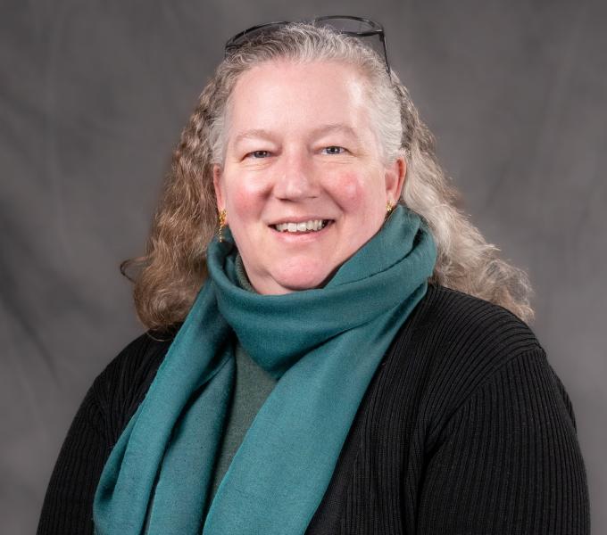 Headshot of a smiling gray-haired woman in a black cardigan and teal scarf.