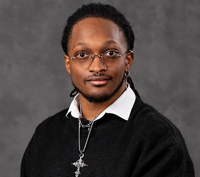 Man in black sweater with white collar sitting in front of gray backdrop.