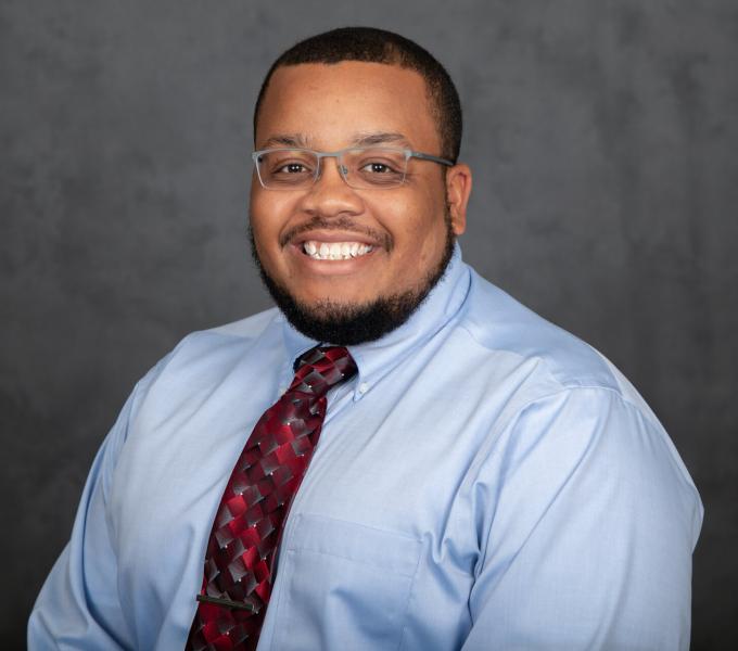 Headshot of smiling man in glasses, a light blue long sleeved shirt, and colorful tie.