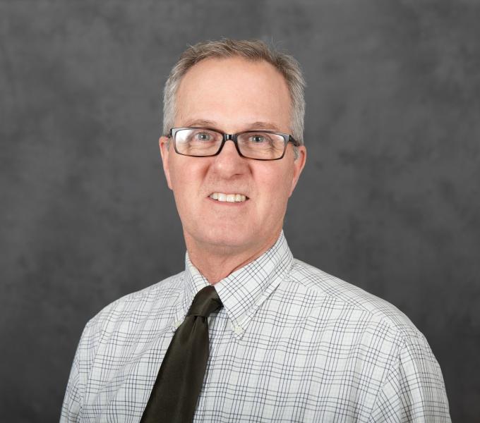 Headshot of a man with gray hair and glasses in a light-colored plaid shirt and dark tie.