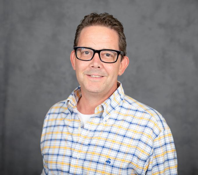 Headshot of man in black plastic glasses and a white, blue, and yellow plaid shirt.