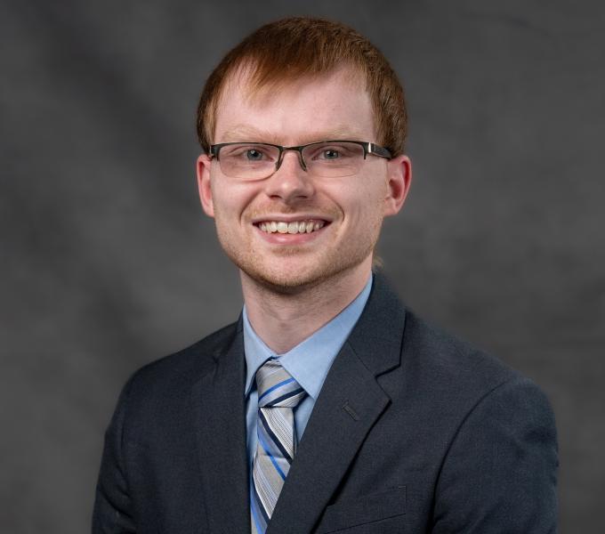 Headshot of a smiling man with short red hair, half-rimmed glasses, and a charcoal-colored suit.