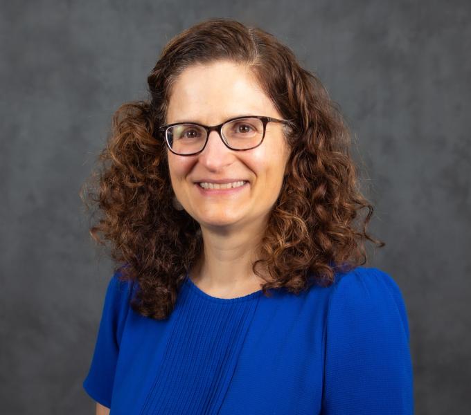 Headshot of a smiling woman in a blue shirt with curly brown hair and glasses.