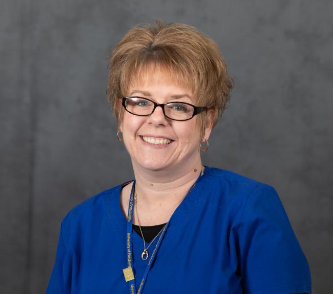 Headshot of smiling woman with short strawberry blonde hair, dark glasses, a blue blouse, and a lanyard.