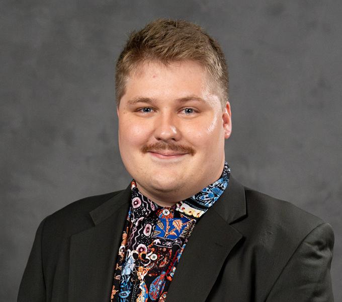 A man with short hair and a mustache smiling in a button up shirt and black jacket.