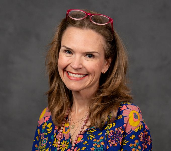 Photo of a woman smiling with long dark hair, red glasses on her head and a blue floral top.