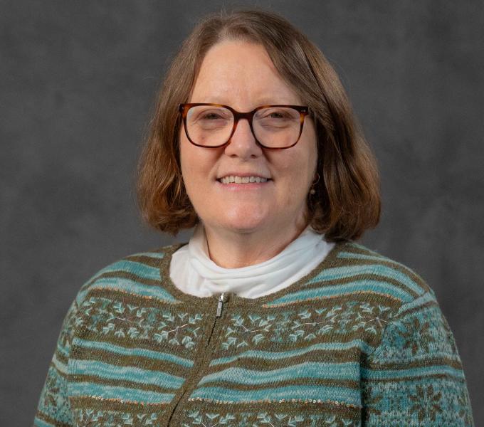 Headshot of a smiling woman with short dark blonde hair in a teal and olive patterned sweater and tortoiseshell glasses.