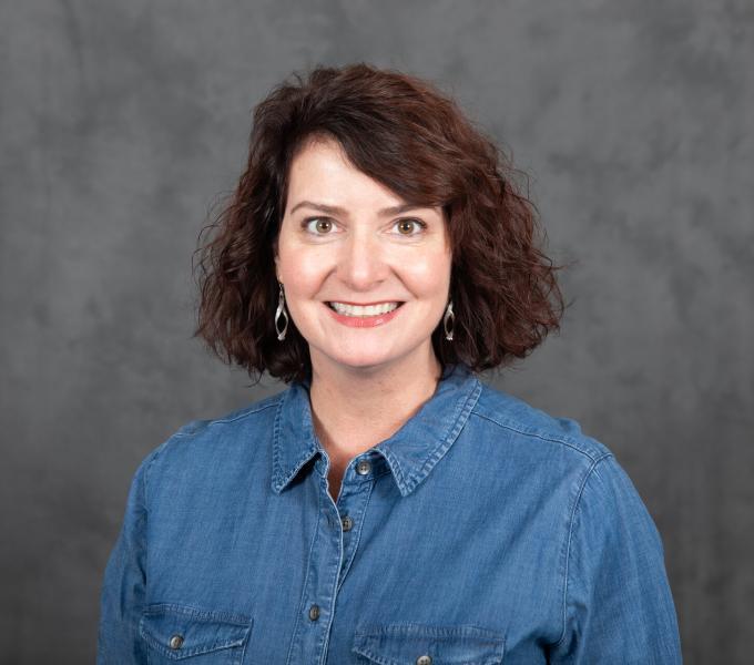 Headshot of smiling woman in a chambray dress with short curly brown hair.