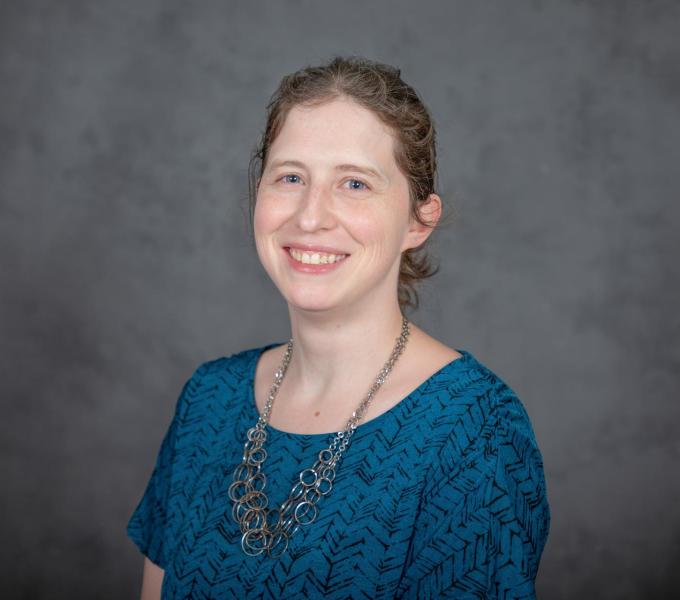 Headshot of smiling woman in a turquoise short-sleeved shirt and large silver ringed necklace.