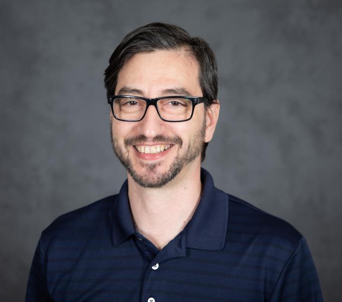 Headshot of smiling bespectacled man with stubble and a dark navy button-down shirt.