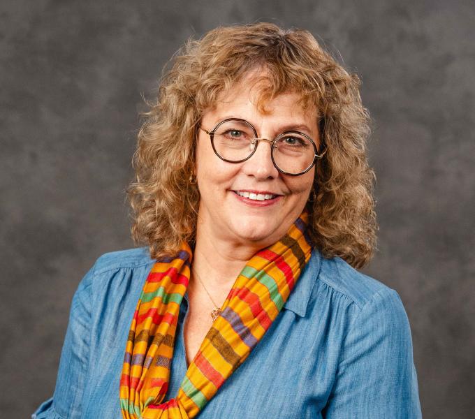 Headshot of a smiling woman with curly golden hair wearing a blue top and an orange scarf.