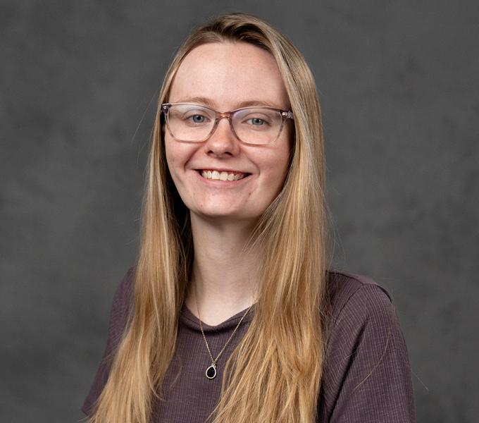 A woman with long blonde hair and glasses smiling in a purple top.