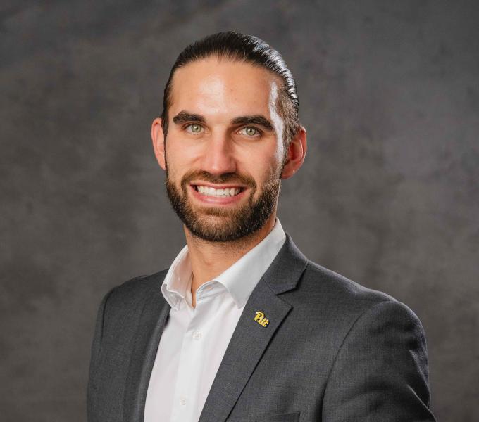 Headshot of a smiling man with slicked back hair and wearing a charcoal sport coat over a white button-down shirt.