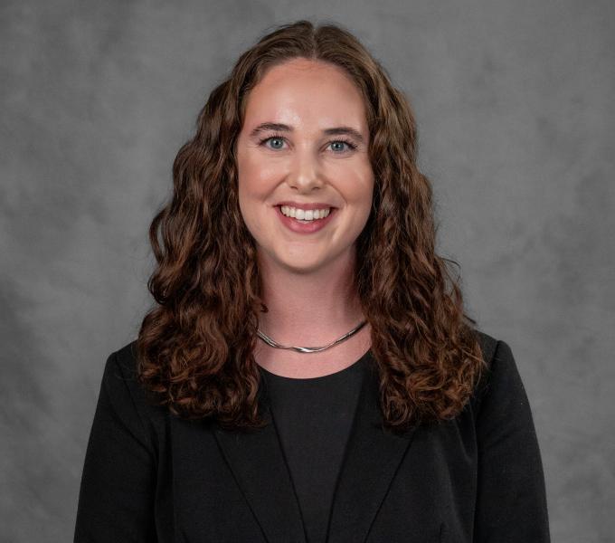 Headshot of a smiling woman with loose curly brown hair in a black blazer and black shirt.