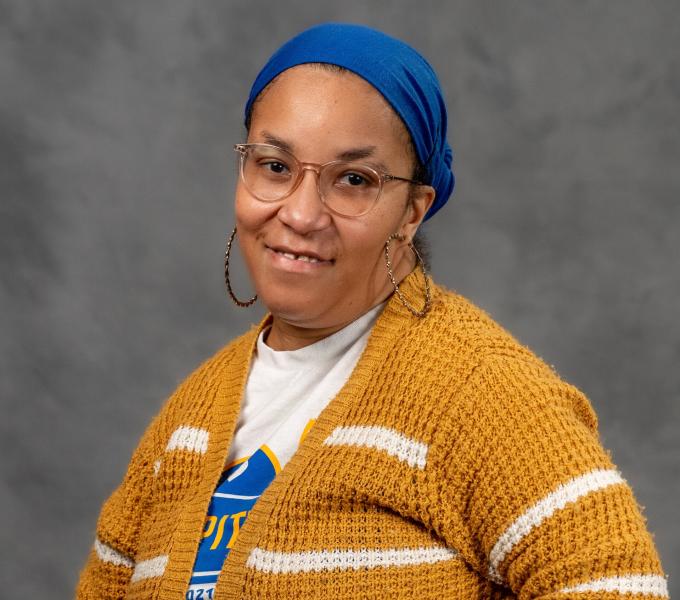 Headshot of a woman in large hoop earrings, a blue hair scarf, gold and white striped sweater, and white tee shirt.