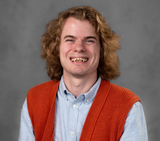 Headshot of a smiling young pan in a light blue button down shirt and orange sweater vest.