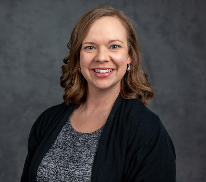 Headshot of smmiling woman with short curled light brown hair in a gray shirt and white cardigan.