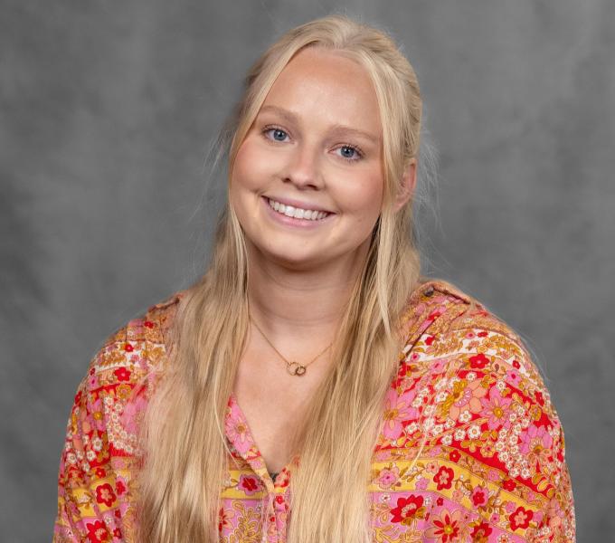 Headshot of a smiling woman with long blonde hair in a pink, yellow, and red floral shirt.