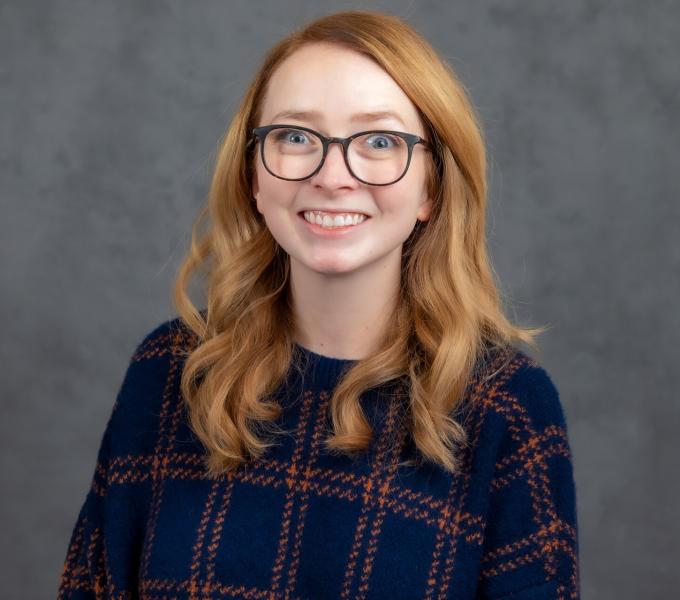 Headshot of smiling strawberry blonde bespectacled young woman in a dark navy plaid shirt.