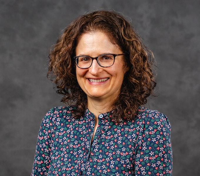 A woman with short brown curly hair and glasses smiling in a blue floral top.