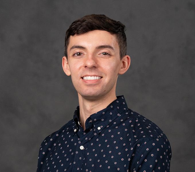 Photo of a man with short dark hair smiling in a navy patterned button up.