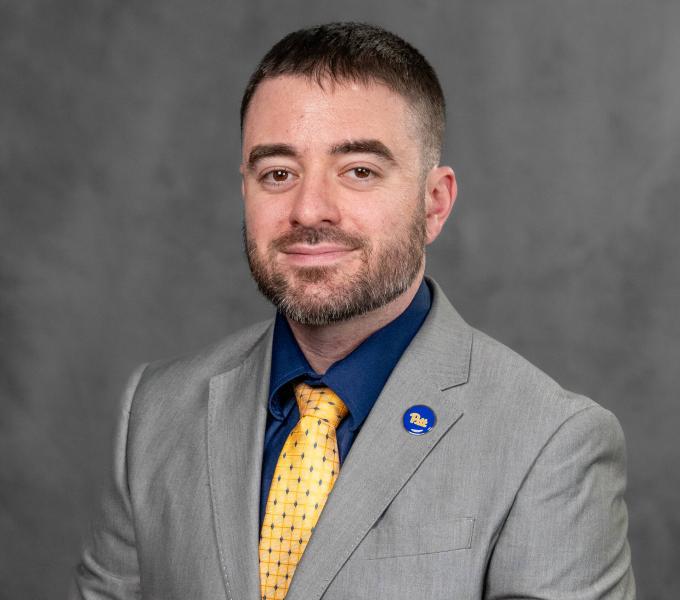 Headshot of a young man in a navy shirt, yellow tie, and gray blazer.