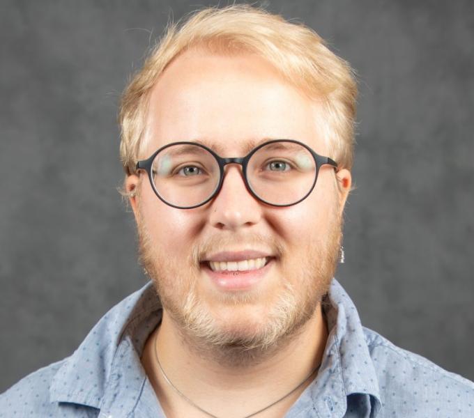 Headshot of a smiling blonde person in glasses and a blue shirt.