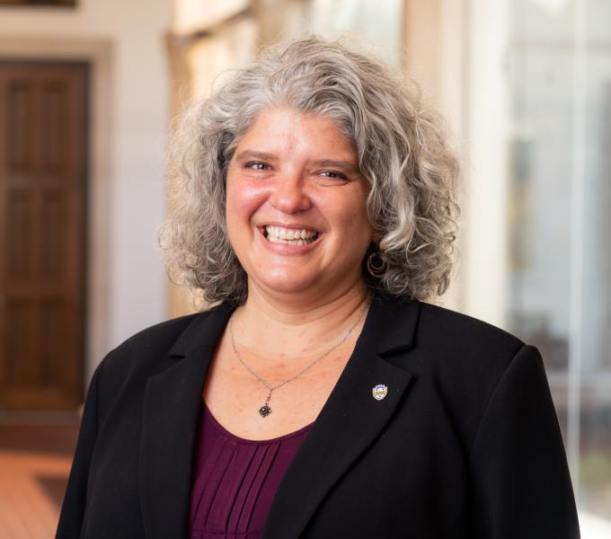 Headshot of a smiling woman with curly gray hair in a black blazer and plum shirt.