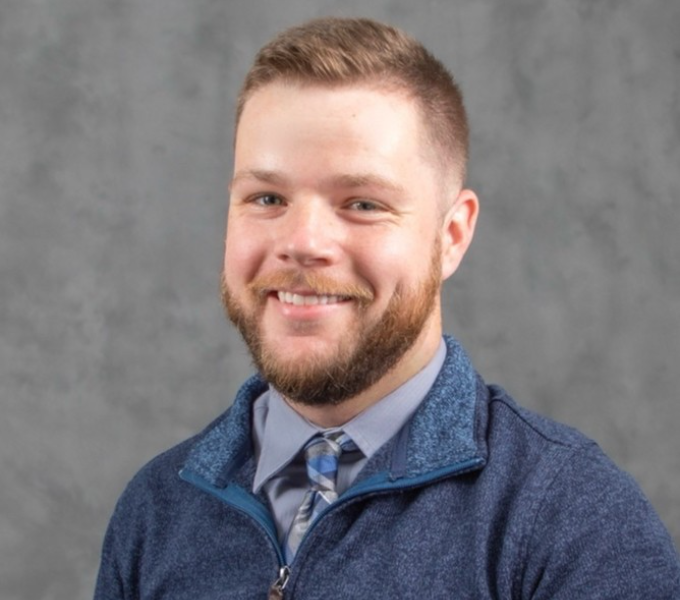 Headshot of smiling man in a gray-blue quarter zip sweater and button down shirt.