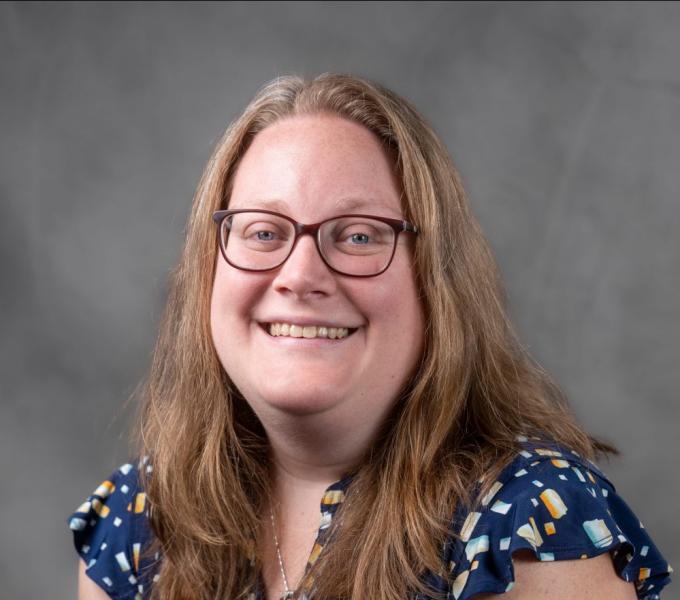 Headshot of a smiling woman with dark blonde hair in glasses and a colorful shirt.