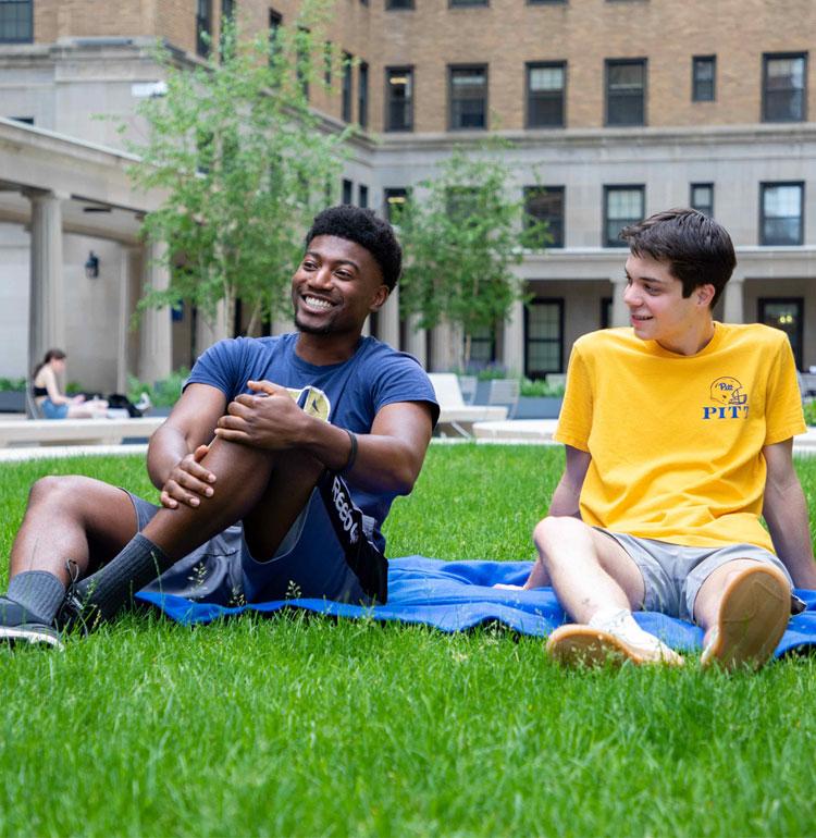 Two young students in Pitt garb sit on a blanket on an outdoor lawn, smiling and talking.